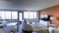 This single-floor living space offers a thoughtfully designed interior that balances modern style with comfortable functionality. The living area serves as the focal point, featuring large floor-to-ceiling windows that flood the space with natural light while framing sweeping city skyline views. A glass door provides direct access to a balcony, further enhancing the connection to the urban landscape outside.

The living room is anchored by a curved white sofa accented with blue throw pillows, paired with a sleek glass-and-chrome coffee table resting atop a warm-toned area rug with flowing abstract patterns. A modern arc floor lamp adds soft ambient lighting, complementing the natural light streaming through the windows. An accent wall finished in a rich terracotta hue introduces warmth and depth to the space, contrasting elegantly with the neutral tones throughout.

The entertainment area features a contemporary white media console supporting a flat-screen television, positioned to be viewed comfortably from the seating arrangement. Dark hardwood flooring runs throughout, grounding the space and adding a refined, polished finish to the overall aesthetic.