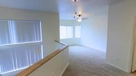 This two-story space is thoughtfully designed to maximize openness and natural light. The upper level, accessible via a staircase with a warm wood-toned railing, reveals a spacious open-plan living area finished with soft, plush wall-to-wall carpeting. Large windows fitted with vertical blinds line the walls, allowing an abundance of natural light to brighten the interior throughout the day. The ceiling fan with a light fixture serves as a functional centerpiece, providing both ventilation and ambient illumination. The smooth, neutral-toned walls complement the light carpeting, creating a clean and airy atmosphere throughout. The open layout offers a generous sense of space, making the upper level feel expansive and versatile. The staircase landing provides a clear sightline across the entire floor, emphasizing the flowing, unobstructed design of the space. Overall, the interior presents a bright, well-lit environment with a neutral color palette that offers a welcoming and adaptable living space across both levels.