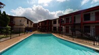 Located on S 11th Street in Kingsville, Texas, this single-story unit is part of a multi-unit residential community defined by its brick and red-accented exterior. The complex centers around a generously sized outdoor swimming pool, enclosed by an iron fence and surrounded by open-sky views — a standout amenity ideal for the warm South Texas climate.

Inside, the unit features consistent tile flooring throughout, offering a clean and low-maintenance surface that complements the cool, light blue-gray painted walls. The open layout connects the main living area to the rest of the unit via a hallway, creating a straightforward and functional flow. Natural light plays a notable role in the interior ambiance, entering through a front window fitted with blinds and, most prominently, through a full-width sliding glass door covered by vertical blinds. This glass door opens to an outdoor patio or walkway with views of the complex's red-trimmed facade, bringing in an abundance of natural light.

The entry is marked by a solid dark-toned door, adding a grounded contrast to the soft interior palette. The unit's design prioritizes simplicity and practicality, with clean white trim, standard HVAC venting, and a neutral finish throughout — offering a functional canvas well-suited for a variety of living arrangements.