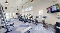 Located in Port St. Lucie, Florida, this fitness center offers a bright, airy workout environment designed to support a full range of exercise routines. The space is defined by its striking vaulted ceilings, which rise dramatically overhead and are complemented by stylish pendant lighting and ceiling fans that keep the atmosphere comfortable throughout the year. Large multi-pane windows line the walls on multiple sides, flooding the interior with natural light and providing views of the surrounding Florida landscape, creating an inviting and energizing ambiance. The flooring features a patterned carpet tile in shades of blue and grey, adding a modern aesthetic while providing underfoot comfort. The cardio section is well-appointed with treadmills, elliptical machines, stair climbers, and smart stationary cycling bikes, each equipped with digital displays and media screens. The strength training area includes a multi-station cable machine, a variety of weight benches, a dumbbell rack, and additional resistance equipment. Full-length mirrors along one wall enhance the sense of space while allowing users to monitor their form. A wall-mounted television is positioned for easy viewing from the cardio equipment. The overall design is clean, modern, and functional, making this fitness center a well-rounded facility for daily workouts.