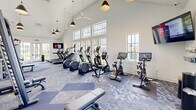 Located in Port St. Lucie, Florida, this fitness center offers a bright, airy workout environment designed to support a full range of exercise routines. The space is defined by its striking vaulted ceilings, which rise dramatically overhead and are complemented by stylish pendant lighting and ceiling fans that keep the atmosphere comfortable throughout the year. Large multi-pane windows line the walls on multiple sides, flooding the interior with natural light and providing views of the surrounding Florida landscape, creating an inviting and energizing ambiance. The flooring features a patterned carpet tile in shades of blue and grey, adding a modern aesthetic while providing underfoot comfort. The cardio section is well-appointed with treadmills, elliptical machines, stair climbers, and smart stationary cycling bikes, each equipped with digital displays and media screens. The strength training area includes a multi-station cable machine, a variety of weight benches, a dumbbell rack, and additional resistance equipment. Full-length mirrors along one wall enhance the sense of space while allowing users to monitor their form. A wall-mounted television is positioned for easy viewing from the cardio equipment. The overall design is clean, modern, and functional, making this fitness center a well-rounded facility for daily workouts.