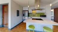 This contemporary single-floor unit is thoughtfully designed with clean lines, modern finishes, and a functional layout visible across its multiple rooms. The kitchen serves as a standout feature, boasting a large central island with an integrated cooktop, a sleek glass range hood suspended from above by pendant lighting, stainless steel appliances including a French door refrigerator and dishwasher, crisp white cabinetry, light stone countertops, and a vibrant green glass tile backsplash that adds a striking pop of color. Cork flooring throughout the kitchen lends warmth and a sustainable touch.

The two bedrooms are each well-appointed with plush dark carpeting and generous windows that fill the rooms with natural light. One bedroom features floor-to-ceiling sliding glass doors offering urban views of the surrounding cityscape, while the other benefits from a tranquil treetop outlook. Both rooms include built-in wooden headboards with integrated shelving and dual reading sconces, alongside ample wardrobe and drawer storage with clean white cabinetry.

A dedicated study nook with a work surface and a large window provides an ideal workspace. The bathroom is finished with large-format gray floor tiles, a walk-in shower with a curtain enclosure, and a narrow frosted window for privacy while maintaining natural light. The overall ambiance is modern, efficient, and bright throughout.