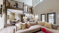 Located at 4901 Tall Timber Dr in Raleigh, NC, this elegantly designed clubhouse serves as a premier amenity space with a layout that is both functional and visually striking. The centerpiece of the space is a dramatic double-height great room, where soaring ceilings and clerestory windows flood the interior with natural light. A mezzanine level with sleek cable-rail balustrades and structural columns adds architectural depth and visual interest overhead.

The open-concept main floor seamlessly integrates a comfortable lounge area, anchored by a large sectional sofa and a stone-accented fireplace with direct access to an outdoor patio through French doors. Adjacent to the lounge, a well-appointed kitchenette features dark cabinetry, a light quartz countertop with an undermount sink, and a tiled backsplash — ideal for entertaining or casual gatherings.

A dedicated dining nook with a round table, modern pendant lighting, and decorative mirror wall art provides an intimate setting. A separate business center offers a built-in workstation, light wood flooring, and ample natural light — perfectly suited for productivity. A cozy sunroom-style sitting area with a built-in window bench and multiple seating options rounds out this versatile and inviting community space.
