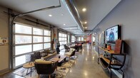 This single-story clubhouse showcases an industrial-chic aesthetic that blends raw architectural elements with comfortable, stylish furnishings. The space is defined by its exposed ductwork running along the ceiling, polished concrete floors, and large garage-style windows that flood the interior with abundant natural light while offering views to the outside streetscape.

The open-plan layout extends generously from front to back, creating a sense of expansive depth. The lounge area at the front features an eclectic mix of woven and upholstered seating arrangements, including armchairs and sofas accessorized with patterned throw pillows, centered around wooden coffee tables atop layered area rugs. This relaxed seating zone transitions seamlessly into a game and entertainment area further within the space, which includes a pool table and additional bar-height seating.

Recessed lighting punctuates the ceiling alongside the industrial ductwork, providing warm illumination throughout. Decorative elements such as vintage signage, wall-mounted maps, and a flat-screen TV add character and personality to the space. The right wall features a long console surface displaying curated décor, enhancing the clubhouse's inviting, community-oriented atmosphere.