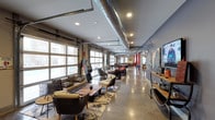 This single-story clubhouse showcases an industrial-chic aesthetic that blends raw architectural elements with comfortable, stylish furnishings. The space is defined by its exposed ductwork running along the ceiling, polished concrete floors, and large garage-style windows that flood the interior with abundant natural light while offering views to the outside streetscape.

The open-plan layout extends generously from front to back, creating a sense of expansive depth. The lounge area at the front features an eclectic mix of woven and upholstered seating arrangements, including armchairs and sofas accessorized with patterned throw pillows, centered around wooden coffee tables atop layered area rugs. This relaxed seating zone transitions seamlessly into a game and entertainment area further within the space, which includes a pool table and additional bar-height seating.

Recessed lighting punctuates the ceiling alongside the industrial ductwork, providing warm illumination throughout. Decorative elements such as vintage signage, wall-mounted maps, and a flat-screen TV add character and personality to the space. The right wall features a long console surface displaying curated décor, enhancing the clubhouse's inviting, community-oriented atmosphere.