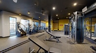 Located at 2125 E Apache Blvd in Tempe, Arizona, this fitness center offers a spacious, single-floor exercise facility designed to support a comprehensive range of workout routines. The space features a striking interior design characterized by dark navy and charcoal ceilings contrasted by bold yellow accent walls, creating an energetic and motivating atmosphere. Rubber flooring throughout provides durability and comfort underfoot across the entire layout.

The cardio zone is positioned along the windowed perimeter, where a row of treadmills, ellipticals, stair climbers, and stationary cycling bikes benefit from natural light and outward views. Large windows along multiple walls enhance the sense of openness and keep the space bright and inviting.

The strength training area is well-appointed with a multi-station cable and weight machine circuit, a dedicated free weights section with a full dumbbell rack, adjustable benches, and a functional training zone equipped with battle ropes and medicine balls. Ceiling-mounted fans and recessed lighting ensure comfortable airflow and consistent illumination throughout.

A built-in shelving unit with yellow cabinetry adds both storage and visual interest near the entrance area, which also features a wall-mounted television for entertainment during workouts. The overall design is modern and purposeful, delivering a fully equipped fitness environment suited for both cardio and strength-focused training.
