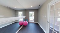 Located at 1 Westchase Dr in Charleston, SC, this single-floor fitness studio is thoughtfully designed to support a variety of exercise and wellness activities. The space features a full-length mirrored wall paired with a mounted horizontal barre, creating an ideal environment for yoga, pilates, dance, or stretching routines. Durable dark rubber flooring provides cushioning and grip underfoot, making it well-suited for both high- and low-impact workouts. Recessed ceiling lights ensure consistent, even illumination throughout the room, while large gridded windows flood the space with natural light and offer a view of the outdoor pool area, enhancing the bright and energizing atmosphere. Neutral warm-gray walls and clean white trim contribute to a calm, focused ambiance. A wall-mounted control panel provides convenient access to climate or lighting adjustments. The open, uncluttered layout allows for flexible arrangement of fitness equipment and accessories, accommodating both individual sessions and small group classes. Overall, this fitness studio combines practical amenities with a welcoming aesthetic to create a well-rounded exercise environment.