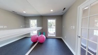 Located at 1 Westchase Dr in Charleston, SC, this single-floor fitness studio is thoughtfully designed to support a variety of exercise and wellness activities. The space features a full-length mirrored wall paired with a mounted horizontal barre, creating an ideal environment for yoga, pilates, dance, or stretching routines. Durable dark rubber flooring provides cushioning and grip underfoot, making it well-suited for both high- and low-impact workouts. Recessed ceiling lights ensure consistent, even illumination throughout the room, while large gridded windows flood the space with natural light and offer a view of the outdoor pool area, enhancing the bright and energizing atmosphere. Neutral warm-gray walls and clean white trim contribute to a calm, focused ambiance. A wall-mounted control panel provides convenient access to climate or lighting adjustments. The open, uncluttered layout allows for flexible arrangement of fitness equipment and accessories, accommodating both individual sessions and small group classes. Overall, this fitness studio combines practical amenities with a welcoming aesthetic to create a well-rounded exercise environment.