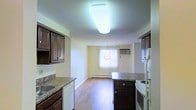 This single-floor residence offers a practical and functional layout centered around an open-plan kitchen and dining area. The kitchen is outfitted with rich, dark wood cabinetry featuring raised panel doors, laminate countertops, and a full suite of white appliances, including a range and dishwasher. A large flush-mounted ceiling light fixture provides bright, even illumination over the kitchen workspace. The space flows seamlessly into a combined dining and living area, where warm hardwood-style flooring runs continuously throughout, lending a cohesive and inviting feel. A central kitchen island with matching dark cabinetry offers additional prep and storage space. The dining area is anchored by a decorative chandelier and benefits from natural light filtering through a window dressed with vertical blinds. A wall-mounted air conditioning unit and baseboard heating ensure year-round comfort. The walls are finished in a soft, neutral tone that keeps the space feeling bright and open. Overall, the layout is well-suited for everyday living, balancing practicality with a warm, understated aesthetic.