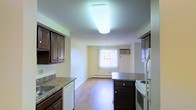 This single-floor residence offers a practical and functional layout centered around an open-plan kitchen and dining area. The kitchen is outfitted with rich, dark wood cabinetry featuring raised panel doors, laminate countertops, and a full suite of white appliances, including a range and dishwasher. A large flush-mounted ceiling light fixture provides bright, even illumination over the kitchen workspace. The space flows seamlessly into a combined dining and living area, where warm hardwood-style flooring runs continuously throughout, lending a cohesive and inviting feel. A central kitchen island with matching dark cabinetry offers additional prep and storage space. The dining area is anchored by a decorative chandelier and benefits from natural light filtering through a window dressed with vertical blinds. A wall-mounted air conditioning unit and baseboard heating ensure year-round comfort. The walls are finished in a soft, neutral tone that keeps the space feeling bright and open. Overall, the layout is well-suited for everyday living, balancing practicality with a warm, understated aesthetic.
