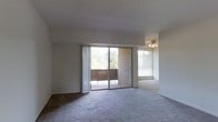 This single-floor residence offers a welcoming and functional layout filled with natural light. The main living area is generously proportioned, featuring soft wall-to-wall carpeting that adds warmth and comfort underfoot. Large windows and a full-width sliding glass door flood the space with daylight and frame lush, tree-filled views, creating a refreshing indoor-outdoor connection. The sliding door opens directly onto a private balcony accented with brick detailing and a wood deck surface, ideal for enjoying the surrounding greenery. A ceiling fan is centrally positioned in an adjacent area, enhancing airflow and adding a practical touch to the overall design. The neutral color palette throughout — from the light walls to the understated flooring — creates a clean, versatile backdrop that adapts easily to a variety of interior styles. The open flow between the main living zone and the adjoining space contributes to an airy, cohesive feel throughout the home. Overall, this space combines everyday functionality with an inviting atmosphere, highlighted by abundant natural light and pleasant outdoor views.