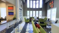 This single-floor lounge space makes an immediate impression with its dramatic double-height ceilings that draw the eye upward toward a curved wall of tall, arched windows. The expansive glazing floods the interior with natural light while offering views of lush greenery outside, creating a bright and inviting atmosphere throughout the day. A contemporary chandelier hangs centrally from the elevated ceiling, adding a refined focal point to the voluminous space.

The main seating area is anchored by plush green velvet sofas and wingback chairs, accented with bold red, navy, and gold throw pillows that introduce vibrant contrast. Seating is arranged in conversational groupings around low-profile tables, encouraging a social and comfortable environment. The flooring features a striking large-scale black and white diamond-pattern tile that adds a classic yet graphic energy to the space.

Along one side of the room, a sleek counter with modern bar-height stools provides an additional seating option, complemented by colorful abstract artwork displayed on the adjacent wall. French-style glass panel doors lead to an adjoining area, contributing to the open, airy character of the space. Overall, the design balances bold color choices with elegant architectural details for a polished, contemporary ambiance.