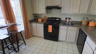 This single-floor space offers a well-organized and functional layout centered around a practical kitchen. The kitchen is equipped with a black electric range and matching overhead ventilation hood, providing a bold contrast against the light-toned cabinetry that lines the walls. The cabinets offer generous storage space both above and below the counters, which feature dark laminate surfaces that provide ample room for food preparation and small appliance placement. The light-colored ceramic tile flooring is easy to maintain and adds a clean, bright quality to the space. Natural light filters in through a window dressed with warm orange curtains, lending a welcoming touch to the room's atmosphere. The countertops are thoughtfully utilized, with space for kitchen essentials and small accessories neatly arranged along the workspace. The overall design is straightforward and practical, prioritizing functionality and ease of use. The cohesive color palette of neutral cabinetry, dark appliances, and light flooring creates a balanced and livable environment suited for everyday cooking and household activity.