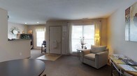 Located at 6785 Oakton Ln in Columbus, Ohio, this two-floor residential space offers a welcoming and functional living environment characterized by soft neutral tones and a comfortable, understated aesthetic. The living area is a highlight of the home, featuring large windows dressed with white blinds and sheer curtains that allow generous amounts of natural light to fill the room. The space is finished with plush wall-to-wall carpeting in a warm beige tone, contributing to an inviting atmosphere. A cozy armchair and accent side table with a small floral arrangement add a homey touch, while a floor lamp provides warm supplemental lighting. The entryway is clearly defined, with a paneled front door accompanied by a small entry mat, creating a natural transition into the main living space. An additional window near the entry further brightens the area. The layout feels open and airy, with thoughtful furniture placement that maximizes usability without overcrowding the room. Subtle architectural details, including a textured ceiling and clean wall lines, lend a classic, timeless character to the interior. Spanning two floors, this space offers room for a variety of living arrangements, making it a practical and comfortable home in a Columbus neighborhood.