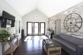 This single-floor community space combines thoughtful design with an inviting, functional layout. The cathedral vaulted ceiling rises dramatically overhead, amplifying the sense of openness and airiness throughout. Rich, dark hardwood-style flooring runs the length of the space, providing a warm and grounding contrast to the light, neutral wall tones.

The most commanding architectural feature is the full-height stacked-stone accent wall, finished in a blend of earthy tones ranging from sandy beige to cool gray. It serves as a striking focal point and anchors a comfortable seating area furnished with a modern gray leather sofa and matching armchair, arranged around a wooden coffee table. A large decorative iron clock mounted on the stone wall adds a bold, industrial touch.

A set of double French doors with divided glass panes floods the interior with natural light and offers views of the outdoor surroundings, creating a seamless visual connection to the exterior. Adjacent to the entrance, a dedicated refreshment station is neatly tucked against the stone wall, equipped with a coffee maker, mini refrigerator, and counter space. A wall-mounted flat-screen television is positioned near a secondary windowed area, adding a media component to the communal setting.

The overall ambiance is warm, modern, and welcoming, blending rustic stone textures with contemporary furnishings to create a comfortable and functional gathering space.