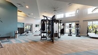 Located at 10 Westedge St in Charleston, South Carolina, this well-appointed fitness center offers a comprehensive workout environment designed to support a wide range of exercise routines. The space is characterized by soaring ceilings with clerestory windows that flood the interior with abundant natural light, creating an open and energizing atmosphere throughout.

The main workout area is generously proportioned and features patterned carpet tiles in neutral and teal tones that define the space with a modern aesthetic. Large floor-to-ceiling windows along one wall provide sweeping views of the outdoor pool area and surrounding landscaping, making cardio sessions more engaging. A bank of treadmills, ellipticals, and stationary bikes is positioned to take full advantage of these views.

The strength training section is equally well-equipped, housing a multi-functional cable and pulley machine, a power rack with an adjustable bench, a full dumbbell rack, and a heavy punching bag. Strategic placement of wall mirrors throughout the space enhances both functionality and the sense of openness. A dedicated area with additional strength machines and benches offers further versatility.

A separate, adjoining room with hardwood-style flooring provides a quieter space suitable for stretching, yoga, or cool-down activities. Ceiling fans, recessed lighting, and a clean, bright color palette of white and teal complete the polished, professional ambiance of this fitness center.