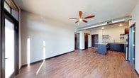 This single-floor space showcases a thoughtfully designed open-concept layout that blends modern and industrial aesthetics. Rich, dark hardwood flooring extends throughout, grounding the space with warmth and contrast against the crisp white walls and ceilings. An exposed metallic ductwork element runs along the upper wall, lending an industrial character that complements the contemporary design. A ceiling fan with wood-toned blades adds both functional airflow and a stylish focal point overhead. The open living area is generously sized, filled with natural light that streams in and casts bright patterns across the floor, enhancing the airy feel of the space. Toward the rear of the unit, the kitchen area features a central island with dark cabinetry, open shelving mounted on the wall, and light upper cabinets — offering a clean and efficient workspace. Dark-framed doors lead to additional areas, including what appears to be a closet with visible hanging space, adding practical storage. Black baseboards provide a sharp, finishing contrast to the light walls. Overall, the space offers a cohesive industrial-modern atmosphere, combining clean lines, quality materials, and an efficient, well-lit open floor plan.