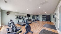 Located at 5 Fall Wood Dr in Hamilton, Ohio, this fitness center offers a well-appointed exercise space designed to accommodate a comprehensive range of workout routines. The room features warm-toned hardwood-style flooring throughout, soft sage-green walls, and a textured ceiling with recessed lighting that keeps the space bright and energizing. Large windows along one side of the room invite natural light and provide outdoor views of the surrounding greenery, creating a refreshing atmosphere for exercise. A wall-mounted ceiling fan adds additional air circulation for comfort during workouts. The cardio section is equipped with multiple treadmills, a recumbent bike, and an elliptical trainer, offering a solid variety of aerobic options. A large wall-length mirror spans one side of the room, visually expanding the space and allowing users to monitor their form. The strength training area includes a multi-functional cable machine, a two-tiered dumbbell rack, and an adjustable workout bench. A dedicated functional training zone houses a well-stocked rack of kettlebells and medicine balls in various weights, alongside a foldable wall-mounted storage unit. A wall-mounted flat-screen television and protective floor mats complete the practical amenities of this comprehensively equipped fitness facility.