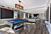 Located at 1600 E Samford Ave in Auburn, Alabama, this single-floor amenity space combines modern farmhouse aesthetics with a welcoming, functional layout ideal for gathering and recreation. Throughout the space, wide-plank hardwood flooring and soft gray shiplap walls create a cohesive, refined atmosphere that balances casual comfort with contemporary style.

The lounge area is generously proportioned, featuring plush neutral seating arranged around a substantial rustic wood coffee table. Multiple windows allow natural light to pour in, enhancing the bright, airy feel of the room. A large wall-mounted flat-screen TV is positioned above a sleek ribbed media cabinet, complemented by warm sconce lighting that adds an inviting ambiance.

Adjacent to the lounge, an open area showcases a striking gallery wall with three bold metallic art pieces mounted above industrial-style bar stools and bistro tables — a perfect spot for casual conversation or small group gatherings.

The recreation zone is anchored by a substantial billiards table with a vivid blue felt surface, suspended beneath a handsome craftsman-style pendant light fixture. A large chalkboard wall and additional bar seating line the perimeter, adding both functionality and character. French doors with glass panels open to an outdoor view, flooding the space with additional natural light and providing easy access to the exterior.