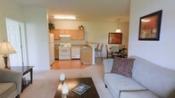 This single-floor residence is designed around an open-concept floor plan that seamlessly connects the kitchen, dining area, and living room into one cohesive living space. The kitchen is outfitted with warm honey-toned wood cabinetry, white appliances including a refrigerator, electric range, and dishwasher, complemented by a flush-mount overhead light fixture. A peninsula with bar-style seating extends from the kitchen, providing a casual dining option and serving as a natural dividing point between the kitchen and the rest of the living space. The flooring in the kitchen is a warm hardwood-style surface, which transitions to plush carpet in the living and dining areas. The dining area accommodates a full table and chair set beneath a pendant light fixture, while a framed piece of artwork anchors the wall. The living room is generously proportioned, featuring neutral-toned carpet and ample space for comfortable seating. The overall ambiance is warm and inviting, with a neutral color palette throughout that creates a bright, cohesive atmosphere well-suited for everyday living.