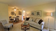 This single-floor space presents a comfortable and cohesive layout, with the living and dining areas flowing naturally into one another. The living area is anchored by a generously sized sofa dressed in neutral tones and accented with patterned throw pillows, complemented by a round wooden coffee table that adds warmth and a classic touch. A pair of framed botanical prints adorns the wall, contributing to the space's understated, tasteful aesthetic. The flooring transitions from plush carpet in the living area to a hardwood-style surface in the dining zone, subtly defining each space while maintaining visual continuity. The dining area accommodates a round table with cross-back chairs, ideal for casual meals or intimate gatherings. A ceiling fan with a built-in light fixture provides both ventilation and ambient illumination throughout. Warm, neutral wall tones lend the space a welcoming glow, while a hallway visible in the background hints at additional rooms beyond. Decorative wall accents, including an oversized clock and architectural panel pieces, add character and personality to the overall design.