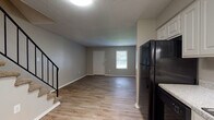 This two-story home offers a practical and well-organized layout across both levels. The main floor features an open living area with wood-style laminate flooring, neutral gray walls, and a textured ceiling. Natural light enters through a front-facing window and the entry door, creating a bright, welcoming atmosphere. The kitchen is positioned adjacent to the living area and is equipped with white cabinetry, light granite-style countertops, a double sink, and black appliances including a refrigerator, dishwasher, electric range, and built-in microwave. A half bathroom is conveniently located on this level, finished with wood-style flooring and neutral tones. A carpeted staircase with a black metal railing leads to the upper floor, where the bedroom is generously sized and features plush carpet, neutral walls, and a set of French doors that allow ample natural light. The bedroom includes two separate closets with shelving and hanging rods, providing substantial storage. A full bathroom is accessible from the upper-level hallway, which also features a window for natural light. The overall design is clean and neutral throughout, offering a move-in-ready environment.