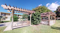 This single-story property is distinguished by its inviting outdoor space, centered around a beautifully crafted wooden pergola. The pergola, constructed from rich, warm-toned timber, features a classic open-lattice roof design that provides partial shade while maintaining an open, airy feel. Horizontal wooden fencing panels complement the pergola's design, adding a modern yet natural aesthetic to the outdoor area.

The grounds are well-maintained, with a neatly trimmed lawn providing a lush green setting. Mature tropical foliage, including palm trees and broad-leafed shrubs, frames the space and contributes to a verdant, tropical ambiance. A secondary pergola structure is partially visible in the background, suggesting the property benefits from multiple dedicated outdoor gathering areas.

The surrounding environment reflects a warm, sunny climate, with the clear blue sky and abundant natural light enhancing the open-air appeal of the space. A neighboring multi-story building is visible in the background, indicating this property is situated within a broader residential community. The combination of the thoughtfully designed wooden structures, manicured landscaping, and tropical plantings creates an outdoor environment that is both functional and visually appealing, well-suited for relaxation and outdoor activities.