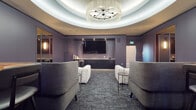 This single-floor amenity space is thoughtfully designed to offer a premium social and entertainment experience. The main lounge area serves as the heart of the space, anchored by a striking circular tray ceiling with integrated ambient lighting and a dramatic sputnik-style crystal chandelier that casts a warm, elegant glow throughout the room. Deep purple-hued walls lend a sophisticated, boutique-hotel atmosphere, complemented by patterned carpet flooring in cool gray tones.

The primary seating area is furnished with generously sized modular sofas and lounge chairs in neutral upholstery, accented by colorful throw pillows and sleek side tables. A large flat-screen television is mounted within a recessed niche along one wall, flanked by built-in dark cabinetry and a beverage refrigerator, creating a polished media wall ideal for gatherings.

Adjacent to the main lounge is a more intimate seating nook, featuring a sectional sofa, accent chairs, and warm pendant lighting beside floor-to-ceiling burgundy drapes. A dedicated golf simulator room provides an immersive experience with a full projection screen and synthetic turf flooring. Nearby, a compact wet bar area with penny-tile backsplash, under-counter refrigeration, and additional lounge seating rounds out the entertainment offerings.