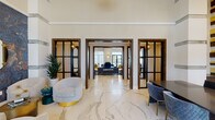 This single-story clubhouse presents a refined, hotel-inspired interior defined by large-format white marble flooring with grey veining that flows seamlessly throughout every zone. The main entrance greets visitors with double-height floor-to-ceiling black-framed glass doors framing views of palm trees, flooding the foyer with natural light. A sculptural reception desk adorned with a striking gold, wood, and marble tile feature wall anchors the entry area alongside steel-framed glass partition doors that lead deeper into the space.

The central lounge area commands attention with soaring ceilings, an artistic geometric metal ceiling installation, and a curated mix of teal, gold, and grey upholstered seating. Bold abstract artwork and wall-mounted televisions enhance the social atmosphere. Adjacent to this is a dedicated bar and lounge featuring a long dark island with bar seating, built-in shelving, and dramatic oversized pendant lighting suspended from double-height ceilings.

A private conference or dining room showcases a textured teal accent wall, gold disc wall art, emerald green seating, and sheer-draped windows with outdoor views. A kitchenette area provides built-in cabinetry, a sink, microwave, and display shelving set against a deep teal backdrop. Throughout, the design balances vibrant color accents with clean architectural lines, creating a dynamic yet cohesive communal environment.