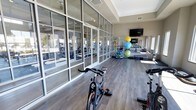 This well-appointed fitness center is designed to provide a bright, open environment for a comprehensive workout experience. The most striking architectural feature is the expansive floor-to-ceiling glass wall system along one side of the space, framed in sleek metal, which floods the interior with natural light and offers views of the surrounding exterior. The high ceilings further enhance the sense of openness and airiness throughout.

The flooring consists of durable hardwood-style planks in a warm gray tone, lending a contemporary aesthetic while remaining practical for high-traffic use. The space is thoughtfully organized to accommodate a range of fitness activities. Stationary cycling bikes are positioned toward the front, making them a focal point upon entry. Along the far wall, a colorful collection of stability and exercise balls is neatly racked alongside additional fitness accessories. A wall-mounted flat-screen television is installed at one end of the room, ideal for guided workouts or entertainment. Mirrored wall panels reflect and amplify the natural light, contributing to the spacious feel. Recessed lighting supplements illumination throughout.