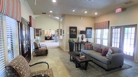 This single-floor space offers an open, inviting layout designed for comfort and functionality. The main living area is anchored by a plush gray sofa accented with rust and earth-toned throw pillows, paired with a rustic wood coffee table and a patterned area rug over stone tile flooring. Warm beige walls and recessed lighting create a bright, welcoming ambiance, while striped Roman shades add a pop of color to the windows that let in generous natural light.

Vaulted ceilings in portions of the space lend an airy, expansive feel, enhancing the sense of openness throughout. Decorative elements such as framed wall art, table lamps, and tasteful accessories contribute to the polished, well-curated aesthetic.

Flowing naturally from the lounge area, the space opens into a work or business center visible in the background, outfitted with computer workstations and seating — ideal for productivity. Additional accent seating, including a patterned armchair, provides flexible arrangements for a variety of uses.

The stone tile flooring transitions seamlessly across the space, unifying the different zones while contributing to the overall warmth and character of the interior. The overall ambiance is casual yet refined, making this a versatile and functional communal environment.