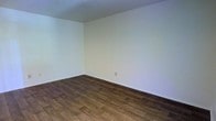 This single-floor space presents a clean, minimalist aesthetic that provides a welcoming blank canvas for a variety of uses. The interior is characterized by crisp, white walls that reflect light evenly throughout the room, contributing to a bright and open atmosphere. Underfoot, rich dark hardwood flooring adds warmth and contrast to the neutral palette, lending a sense of quality and durability to the space. The flooring features a classic plank layout with natural wood grain variations, adding visual texture and depth. The room's proportions feel comfortable and well-balanced, with smooth white ceilings that complement the simplicity of the walls. A standard electrical outlet is positioned along the baseboard, hinting at practical utility within the uncluttered design. The white baseboards provide a clean transition between wall and floor, reinforcing the neat, well-maintained character of the space. Overall, the interior exudes a straightforward, functional ambiance — ideal for those seeking a move-in-ready environment that can be easily adapted to suit a range of living or working needs.