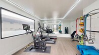 Located at 3454 Ruffin Rd in San Diego, California, this amenity space combines a well-equipped exercise room with a vibrant outdoor courtyard designed for both activity and relaxation. The fitness room features clean white walls, light wood-style flooring, and recessed perimeter lighting that lends a modern, polished aesthetic. It is outfitted with a treadmill, elliptical, and stationary bike — including a screen-equipped cycle — all positioned on protective floor mats. A wall-mounted air conditioning unit ensures year-round comfort, while a window provides a view to the exterior. Adjacent to the fitness room, the outdoor courtyard serves as a social hub. Artificial turf creates a lush, low-maintenance green space underfoot, while a shade sail and string lights overhead add ambiance and protection from the sun. The space is divided into distinct zones: a lounge area with cushioned chairs and a coffee table enclosed by sleek dark horizontal slat fencing, and a BBQ station featuring dual built-in grills set against stacked stone, topped with a decorative metal canopy. A round dining table with modern chairs completes the al fresco setup, making this courtyard a versatile gathering space for outdoor dining and leisure.
