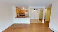 This single-floor unit is designed with a practical, open-concept layout that promotes a seamless flow between living areas. Light oak hardwood flooring runs throughout the main living spaces, lending warmth and continuity to the interior. The living area is generously proportioned, with neutral cream-toned walls, a wall-mounted air conditioning unit, and access to a private balcony through a sliding glass door, allowing natural light to filter into the space.

The kitchen is positioned adjacent to the main living area and is separated by a peninsula counter finished with a granite-style surface and a double stainless steel sink. Light wood cabinetry lines the walls, providing ample storage, while white appliances — including a gas range and a full-size refrigerator — complete the functional layout. A flush-mount ceiling fixture illuminates the kitchen area.

A ceiling fan is installed in the dining or secondary living zone, enhancing air circulation. The bedroom spaces feature the same hardwood flooring and neutral walls, with baseboard heating units installed along the perimeter for climate control. Windows allow natural light and outdoor views into the bedrooms. The bathroom includes a full-size tub and shower combination with chrome fixtures, a toilet, and clean white finishes throughout.