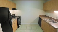This single-floor space features a kitchen designed for practical use, with a clean and straightforward layout. The kitchen is equipped with black appliances, including an electric coil range with an overhead exhaust hood and a dishwasher, seamlessly integrated alongside light wood cabinetry. The cabinetry provides ample storage both above and below the countertops, which feature a granite-style laminate surface. A sink is positioned on the right side of the kitchen, complementing the functional workflow of the space. The light beige tile flooring runs throughout the kitchen, offering a neutral and easy-to-maintain surface. The walls are painted in a soft, neutral tone, contributing to a bright and open feel. White baseboards provide a clean contrast against the flooring and walls. Overall, the kitchen presents a utilitarian design that prioritizes functionality, making efficient use of the available space with clearly defined cooking, preparation, and cleaning zones.