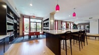 This single-floor library is thoughtfully designed to balance functionality with a warm, inviting ambiance. The open layout is anchored by a substantial dark-wood central island with a light stone countertop and bar-height seating, serving as a versatile gathering or work surface. Vivid magenta pendant lights hang overhead, adding a bold contemporary accent against the neutral ceiling.

Extensive floor-to-ceiling dark bookshelves line multiple walls throughout the space, housing a rich collection of books and media. A circular upholstered bench provides a central seating focal point, complemented by plush armchairs placed in reading nooks near the shelving. Bamboo hardwood flooring runs throughout, contrasting with area rugs that define specific zones.

Large floor-to-ceiling windows with deep red curtain panels allow generous natural light and offer outdoor views, brightening the dining-style reading table surrounded by red chairs. A separate enclosed room—accessible through glass-paneled doors—features an exposed brick accent wall, a large conference table, multiple computer workstations, bold artwork, and a pendant light fixture, making it ideal for private study or group work. The overall atmosphere combines modern design elements with a classic library aesthetic.