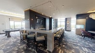 Located at 44 Maryland Ave in Rockville, Maryland, this elegantly designed single-floor amenity space offers a seamless blend of modern aesthetics and functional versatility. The layout encompasses several distinct zones connected by carpeted corridors adorned with curated artwork and sleek dark-wood accent walls with contemporary sconce lighting.

At the heart of the space is a dramatic bar area featuring an expansive white countertop island surrounded by high-back bar seating. Overhead, a striking geometric pendant lighting system adds an architectural focal point, while a wall-mounted flat-screen TV and mosaic tile backsplash lend a polished, upscale ambiance.

The adjacent kitchen is fully equipped with dark cabinetry, white quartz countertops, stainless steel appliances, beverage refrigerators, a sink, and warm cove lighting — ideal for catering or event preparation.

A cozy lounge seating area features plush lavender swivel chairs, marble-top side tables, and a sculptural woven pendant light. Vibrant abstract artwork and a dark feature wall with a recessed display create a gallery-like atmosphere. Floor-to-ceiling windows in this area offer sweeping elevated views of the Rockville skyline and surrounding treetops, flooding the space with natural light.

Dark-framed glass entry doors, patterned carpeting throughout, and a refined, cohesive design language unify this upscale communal space.