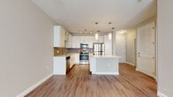 Located at 5671 Mauna Loa Blvd in Sarasota, Florida, this single-story residence offers a modern, open-concept layout designed for comfortable everyday living. The heart of the home is a well-appointed kitchen featuring crisp white cabinetry, a tasteful tile backsplash, and a full suite of stainless steel appliances including a range, microwave, refrigerator, and dishwasher. A generously sized center island with a light countertop provides additional prep space and doubles as a casual gathering spot, complemented by stylish pendant lighting overhead. Track lighting adds further illumination throughout the open living area, which is finished with warm wood-look flooring that extends seamlessly across the main living spaces. A ceiling fan enhances comfort in the living area, which benefits from natural light through a window with blinds. The bathroom is finished with neutral tile flooring, white cabinetry with stone-look countertops, and a glass-enclosed shower, maintaining the clean, contemporary aesthetic found throughout the home. A dedicated laundry area houses a full-size top-loading washer and dryer with overhead shelving for added storage convenience. The overall design palette of soft neutrals, white finishes, and warm wood tones creates a bright, cohesive, and inviting atmosphere throughout.