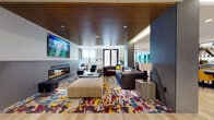 This thoughtfully designed single-floor amenity space combines style and functionality across several distinct yet interconnected zones. Light hardwood flooring runs throughout, providing a warm, cohesive foundation that ties together the varied areas.

The centerpiece lounge area makes an immediate impression with its rich wood slat ceiling, a wall-mounted flat-screen television, and a sleek linear fireplace. Plush sectional seating is anchored by a bold, multicolored abstract area rug, creating an inviting and energetic atmosphere for relaxation and social gathering.

Adjacent to the lounge is an open dining area furnished with a large dark-topped communal table surrounded by vibrant yellow upholstered chairs, lit by a striking geometric teal pendant light. Colorful original artwork adorns the walls, adding artistic character to the space.

The kitchenette features dark cabinetry, stainless steel appliances, a subway tile backsplash, and distinctive V-shaped open shelving with under-cabinet lighting, offering both practicality and visual interest.

Flexible banquette seating with teal accent walls provides additional casual gathering spots, while glass barn doors open to dedicated workspaces and meeting areas with teal partition walls. Recessed lighting and roller shades throughout ensure a bright, comfortable environment adaptable to a variety of uses.