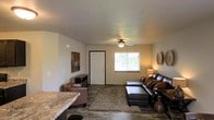 This two-floor residential space offers a comfortable and functional layout designed for everyday living. The main living area is immediately welcoming, featuring dark hardwood-style flooring that contrasts elegantly with the warm, neutral-toned walls. A ceiling fan centered overhead provides both practical ventilation and a subtle design accent to the room. Natural light filters in through a window near the front entry door, brightening the space and creating an airy atmosphere. The living room provides ample space for seating and relaxation, complemented by a plush area rug that adds warmth and definition to the room. A granite-style countertop is visible at the edge of the living area, suggesting a connected kitchen or breakfast bar that contributes to the open-concept feel of the main floor. Wall art and decorative accents add personality and a lived-in charm to the space. The layout flows naturally, making it easy to navigate between functional zones. With two floors, the residence provides separated living and private spaces, offering a practical division between communal and personal areas. Overall, the space presents a cozy, well-appointed environment with a neutral palette and durable finishes suited to comfortable daily living.