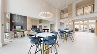 This expansive single-floor amenity and clubhouse space is designed to impress, offering a generous footprint organized into several distinct yet interconnected zones. The architecture is defined by dramatic double-height ceilings throughout much of the space, with clerestory windows and floor-to-ceiling glazing that flood the interiors with abundant natural light and provide views of the surrounding landscape.

The main lounge serves as the centerpiece, anchored by two sculptural brass branch-style chandeliers suspended over pairs of teal sofas arranged around a marble-top coffee table. Polished large-format marble-look tile flooring runs throughout, lending a refined, upscale character. A wide corridor with light wood accent columns and a mezzanine-level overlook connects the lounge to the entry vestibule, which features a dramatic two-story window wall.

Adjacent areas include a game room with a pool table and foosball table, multiple intimate seating nooks with built-in wall-mounted displays, and a quiet lounge adorned with abstract artwork. A dedicated café area offers a coffee and beverage bar with warm cabinetry and a beverage cooler. Bar-height communal tables with modern seating provide additional gathering options throughout the space.