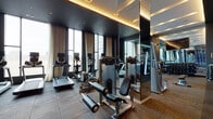 This upscale fitness center combines thoughtful design with comprehensive workout offerings in a single-floor layout. Entry is through striking black-framed glass partition doors, setting a refined tone from the outset. The interior is defined by a dark ceiling with warm cove lighting and recessed spotlights, creating a polished, moody ambiance throughout.

The main workout area is spaciously laid out with rubber-tiled flooring and features a broad selection of strength training equipment, including cable machines, weight benches, and a full dumbbell rack along one mirrored wall. Full-length mirrors run the length of one side, visually expanding the space and aiding form during exercise. On the opposite wall, an eye-catching large-scale mural depicting athletic figures in motion adds a dynamic, artistic focal point.

The cardio section is positioned along floor-to-ceiling windows dressed with sheer curtains, providing natural light and outward views. Treadmills, ellipticals, and stationary bikes are neatly arranged in this zone. A reception-style counter with a granite top, metallic mosaic tile backsplash, and dark cabinetry with open shelving serves as a welcoming amenity station. Together, these elements create a fitness environment that is both functional and visually refined.