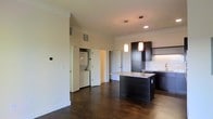 This single-floor space showcases a thoughtfully designed open-concept layout that seamlessly integrates the kitchen and living areas. The kitchen is anchored by a central island with a granite countertop, providing both prep space and a natural gathering point. Dark espresso cabinetry lines the kitchen walls, complemented by under-cabinet lighting and a clean white tile backsplash. Two pendant lights hang above the island, adding focused illumination and a contemporary aesthetic touch. Additional recessed lighting ensures the space remains well-lit throughout.

Rich, dark hardwood flooring flows through the main living area, transitioning to lighter flooring near the kitchen, creating a subtle visual distinction between the two zones. The ceilings are generously elevated, contributing to an airy and open feel. A dedicated in-unit laundry area is conveniently tucked behind a doorway, housing a stacked washer and dryer. Neutral wall tones throughout keep the interior feeling clean and versatile. The overall ambiance is modern and functional, with a cohesive dark-and-light contrast defining the design character of the space.