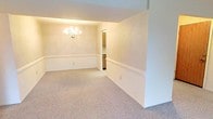 This single-story space offers a straightforward, functional layout connecting a formal dining area to a kitchen. The dining room is finished with neutral wall-to-wall carpeting and warm, cream-toned walls accented by chair rail molding that runs along the perimeter. A traditional multi-arm chandelier serves as the room's focal point, providing ambient lighting overhead. An open passthrough cut into the dividing wall creates a convenient visual and physical connection between the dining space and the kitchen beyond.

The kitchen features a practical U-shaped layout with ample storage provided by medium-toned wood cabinetry spanning both the upper and lower sections of the walls. Light-colored laminate countertops offer generous prep space, and a double-basin stainless steel sink is positioned along one wall. A freestanding electric coil range with an overhead ventilation hood serves as the cooking centerpiece. Light-toned tile flooring runs throughout the kitchen, contrasting with the carpeted dining area. Together, the spaces present a classic, no-frills interior well-suited for everyday living.