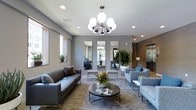 This single-floor space showcases a thoughtfully designed lounge or common area with a distinctly modern aesthetic. The room is anchored by a striking sputnik-style chandelier with globe bulbs, serving as a focal point that draws the eye upward toward the smooth gray ceiling, which is also equipped with recessed lighting for layered illumination throughout the space.

The seating area features a sleek gray leather sofa paired with coordinating blue-gray upholstered armchairs, all arranged around a round, dark metal coffee table. A textured area rug in neutral tones grounds the furniture grouping and adds warmth to the overall palette. Fresh floral and tropical plant arrangements contribute vibrant pops of color against the cool gray walls.

Three tall, narrow mirrors mounted along the back wall create a sense of depth and reflect natural light that filters in through windows fitted with roller shades. Additional tropical potted plants flank the mirrors, enhancing the fresh, inviting atmosphere. An elevator and corridor are visible toward the rear, suggesting this functions as a communal reception or lobby area. The overall ambiance is polished, contemporary, and welcoming.