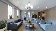 This single-floor space showcases a thoughtfully designed lounge or common area with a distinctly modern aesthetic. The room is anchored by a striking sputnik-style chandelier with globe bulbs, serving as a focal point that draws the eye upward toward the smooth gray ceiling, which is also equipped with recessed lighting for layered illumination throughout the space.

The seating area features a sleek gray leather sofa paired with coordinating blue-gray upholstered armchairs, all arranged around a round, dark metal coffee table. A textured area rug in neutral tones grounds the furniture grouping and adds warmth to the overall palette. Fresh floral and tropical plant arrangements contribute vibrant pops of color against the cool gray walls.

Three tall, narrow mirrors mounted along the back wall create a sense of depth and reflect natural light that filters in through windows fitted with roller shades. Additional tropical potted plants flank the mirrors, enhancing the fresh, inviting atmosphere. An elevator and corridor are visible toward the rear, suggesting this functions as a communal reception or lobby area. The overall ambiance is polished, contemporary, and welcoming.