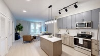 Located at Kenilworth Terrace NE in Washington, DC, this thoughtfully designed one-bedroom, two-bathroom residence offers a sleek, modern aesthetic throughout its efficiently utilized single-floor layout. The open-concept kitchen and living area serves as the heart of the home, anchored by a central island with a crisp white countertop and warm wood base. The kitchen is outfitted with gray upper cabinetry, light wood lower cabinets, a classic white subway tile backsplash, stainless steel appliances—including a dishwasher, range, and microwave—and a matte black faucet, striking a refined contrast against the light finishes. A pendant and track lighting combination adds both function and modern flair. The living area flows naturally from the kitchen, bathed in natural light from large windows with views of the surrounding neighborhood. Light wood-toned flooring runs throughout, unifying the space with warmth and continuity. The bedroom offers a cozy, well-proportioned retreat with a window framing neighborhood views. Both bathrooms are finished with large-format tile, a soaking tub, and light wood vanities paired with matte black hardware, delivering a clean and contemporary feel throughout.
