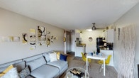 This single-floor space is designed with an open-concept layout that seamlessly connects the living area, dining space, and kitchen into one cohesive and functional environment. The living room features a comfortable gray sofa accented with decorative pillows in blue and yellow, complemented by a glass coffee table and a set of bold abstract wall art pieces that add visual interest to the neutral-toned walls. Plush carpet underfoot provides warmth and comfort throughout the living area. The dining space sits adjacent to the living room and is furnished with a clean white table and vibrant yellow chairs, creating a lively and inviting atmosphere. A ceiling fan overhead provides both light and airflow to the area. The kitchen is efficiently laid out with white cabinetry, a dark tiled backsplash, and black appliances including a refrigerator and range. A single window above the sink allows for natural light to filter into the cooking area. Potted greenery on top of the cabinetry adds a touch of nature to the space. The unit's textured ceilings and consistent neutral wall tones provide a clean, understated backdrop that allows the bright accent colors and furnishings to stand out throughout the space.