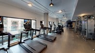 Located at 110 N Myers St in Oceanside, CA, this well-appointed fitness center offers a comprehensive workout environment across a single, elongated floor plan. The space is designed to accommodate a wide range of exercise needs, thoughtfully divided into distinct zones for cardio, strength training, and functional fitness.

The cardio section is lined with a robust row of Life Fitness treadmills, elliptical trainers, and stair climbers, positioned alongside large floor-to-ceiling windows that allow generous natural light and views of the surrounding streetscape. Ceiling fans and recessed lighting complement the bright, airy atmosphere throughout.

The strength training area features a full rack of dumbbells displayed along mirrored walls, adjustable benches, cable machines, and a dedicated TRX suspension training station. Bold teal and navy accent walls add a dynamic visual character to the space, contrasting with the neutral wood-toned flooring and concrete structural columns.

Additional amenities include wall-mounted flat-screen televisions positioned for cardio users, a water fountain station, and restroom access. The open layout and wide central corridor ensure smooth circulation between zones, making the facility functional and accessible. The industrial-style exposed ductwork and ceiling details lend the space a contemporary, energetic ambiance suited to an active fitness environment.