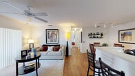 This single-floor residence offers a thoughtfully designed layout that seamlessly connects its main living areas. The living room serves as the heart of the home, featuring warm hardwood-style flooring, a ceiling fan, and a large sliding glass door fitted with vertical blinds that allow natural light to filter in while providing access to an outdoor patio. A plush area rug anchors the seating area, complemented by tasteful wall art and a tall decorative floor lamp.

Adjacent to the living room, the dining area accommodates a glass-top table with wood chairs and is accented by floating wall shelves displaying decorative accessories. The kitchen is efficiently laid out in a U-shape configuration, offering warm honey-toned cabinetry, ample counter space, a black range, refrigerator, and dishwasher, all set atop hardwood-style flooring.

The primary bedroom is generously sized with soft carpeting, dual nightstands, and two large windows with vertical blinds that bring in natural light. A second bedroom mirrors this comfortable style. The full bathroom features a wide vanity with a large mirror, extended countertop, under-sink storage, and a glass-enclosed shower, all finished with hardwood-style flooring for a clean, cohesive look throughout.